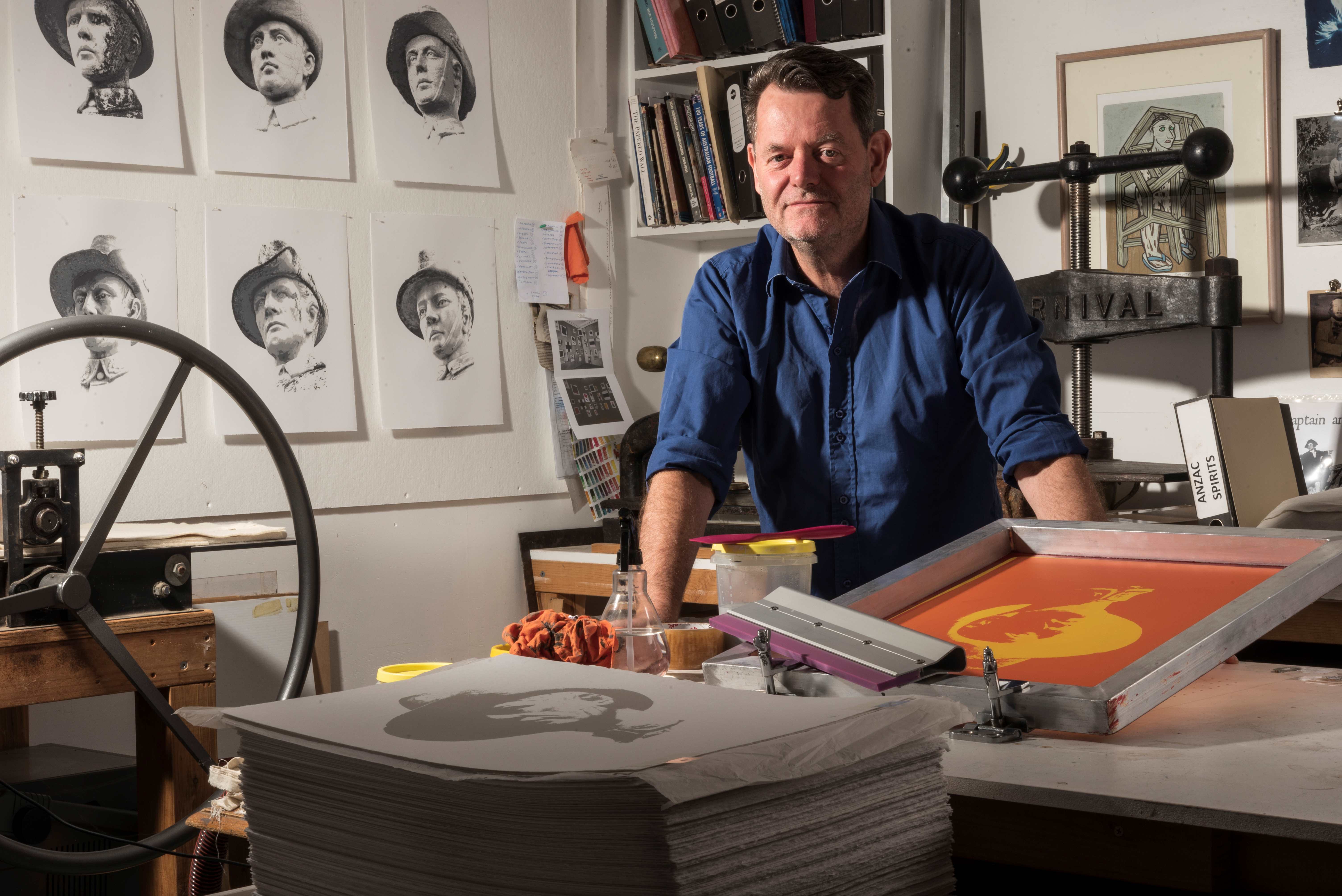 The image shows an artist standing in a studio, surrounded by prints of soldier statues on the wall behind him. He is wearing a blue shirt and leaning on a table with stacks of prints and screen printing tools. The workspace is filled with art supplies, books, and equipment,.