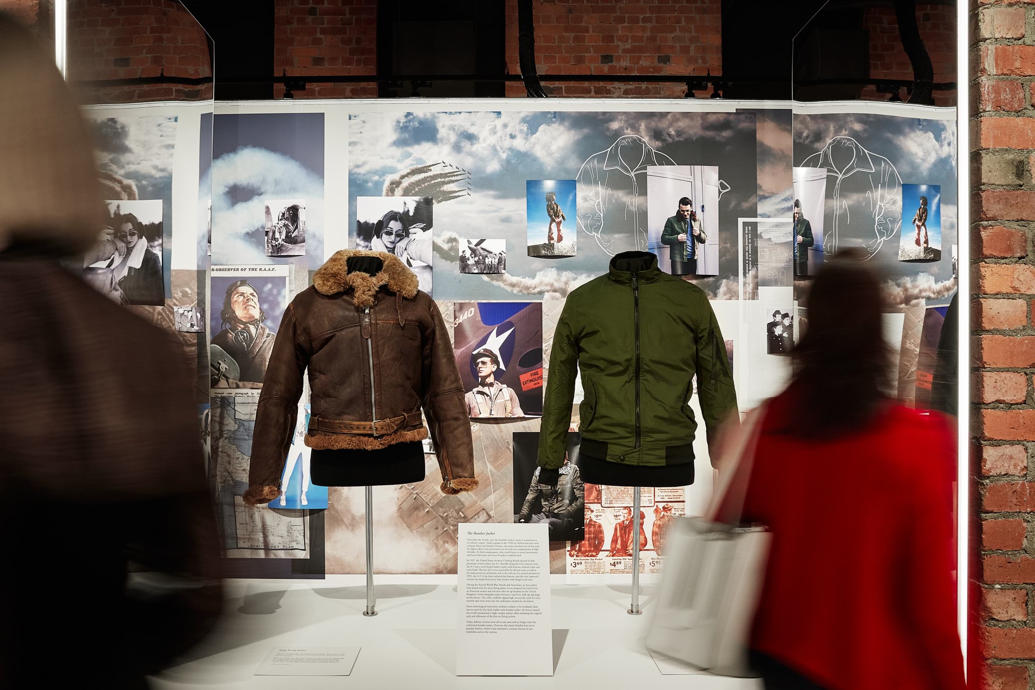 In the centre of this image are two bomber jackets. The one on the left is one from the war with brown leather and fur lining. The one on the right is green with a black zipper, and is a modern fashion version of the jacket. Behind the jackets there is a wall with collaged images of pilots. In the foreground are the blurred images of two women walking past the jackets. 