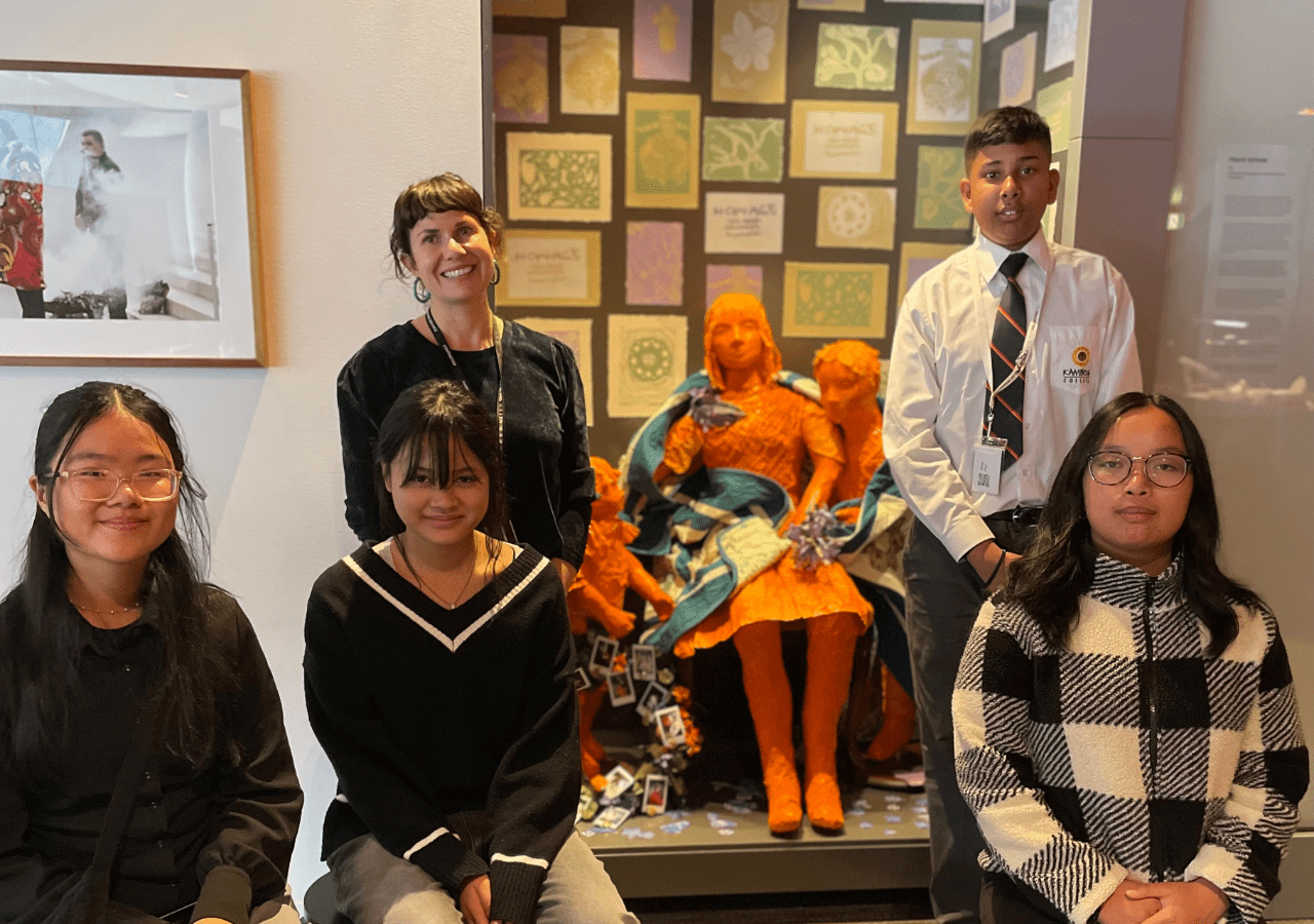 Group of Students sitting and standing in front of an artwork they have created as a message of remembrance and reflection. The artwork is 3 scupltures showing a mother, son and daughter surrounded by lino prints of symbols of remembrance.
