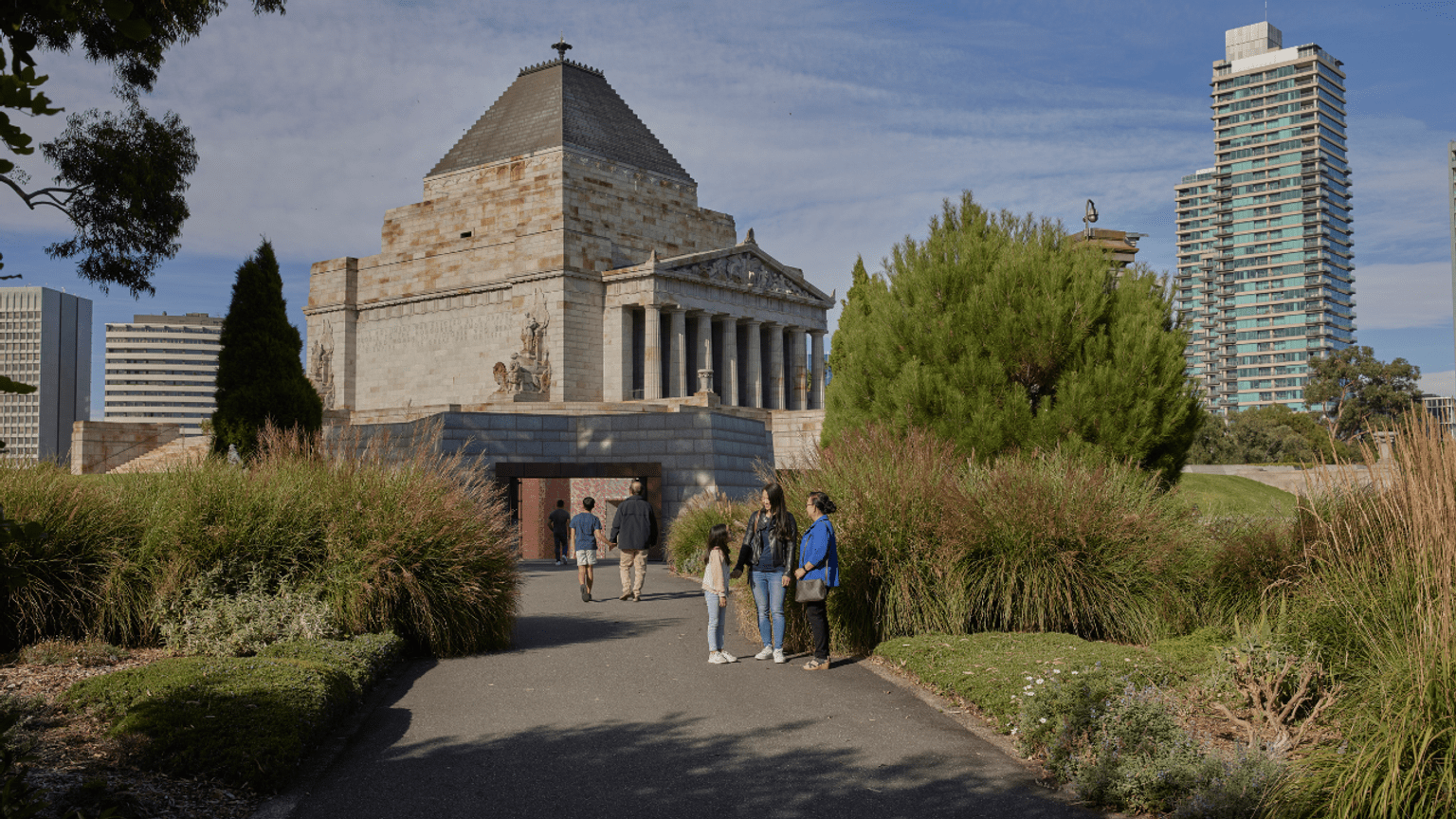 Family standing on a garden path with green plans and trees. The Shrine of Remembrance, a classical building is in the background.